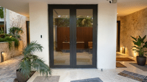 A wide-angle view of a luxury home entrance featuring A&J Windowhaus dark bronze French doors, travertine stone walls, and lush tropical landscaping.