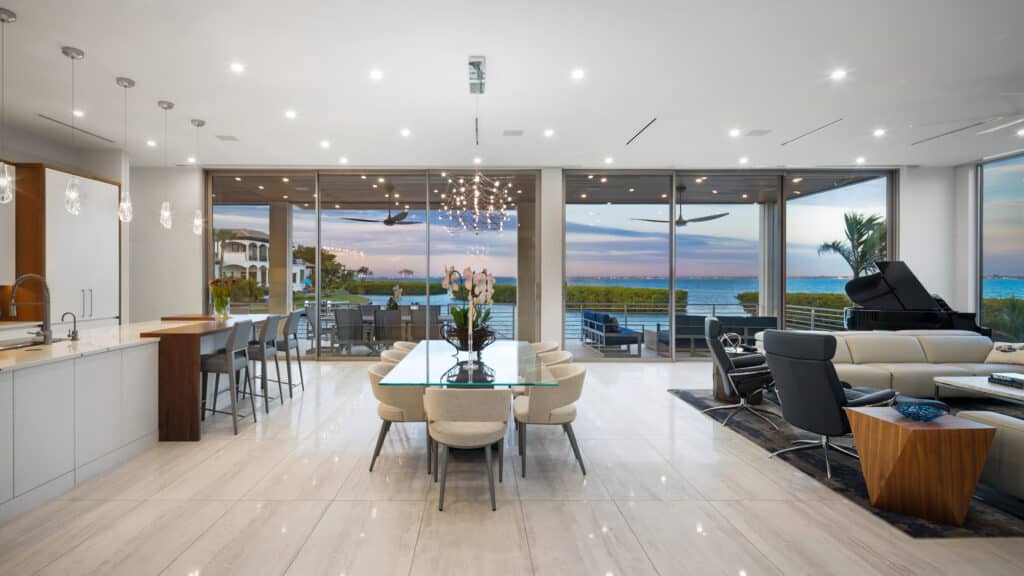 An elegant modern open-plan home interior features a large wall of impact-resistant sliding glass doors, showcasing the type of extensive project achievable with professional Storm Window Replacement, overlooking a tranquil coastal bay with palm trees at dusk.