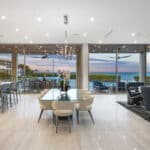 An elegant modern open-plan home interior features a large wall of impact-resistant sliding glass doors, showcasing the type of extensive project achievable with professional Storm Window Replacement, overlooking a tranquil coastal bay with palm trees at dusk.