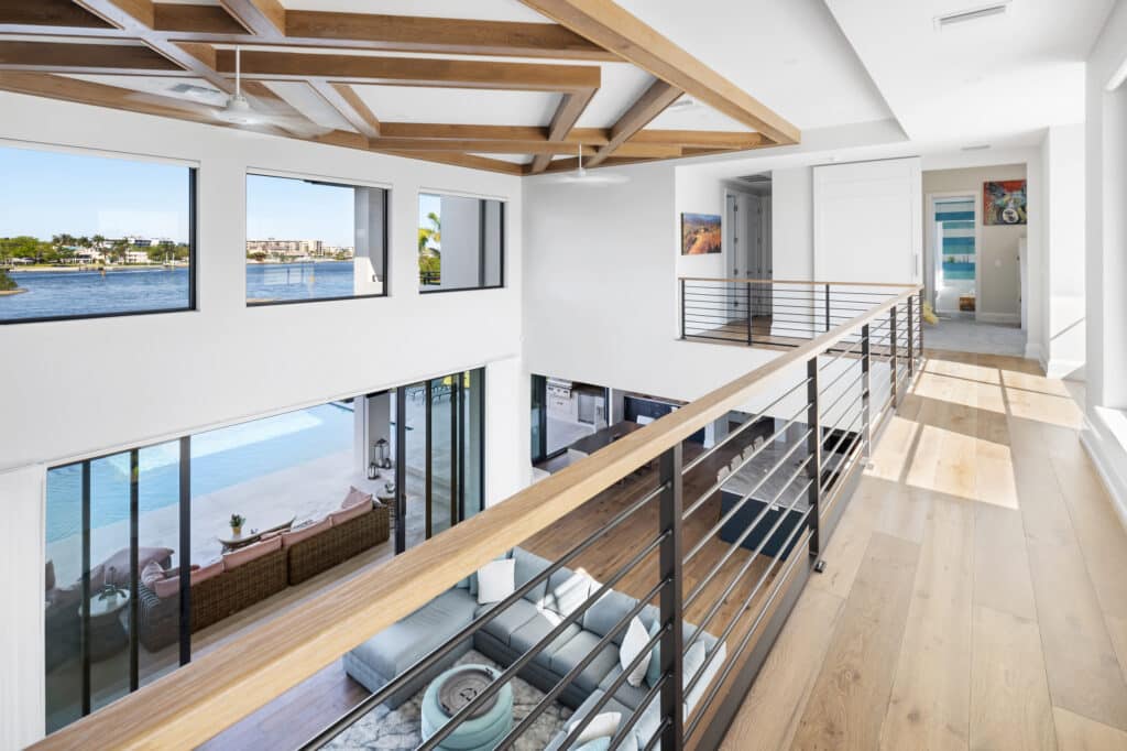 A second-story walkway overlooks a modern living area, where large windows and a sliding door framing a scenic coastal waterway highlight a premium Storm Window Replacement.