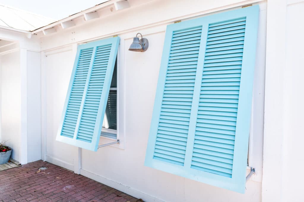 Light blue coastal Bahama shutters propped open on a white home exterior, providing stylish Storm Windows Hurricane Protection.