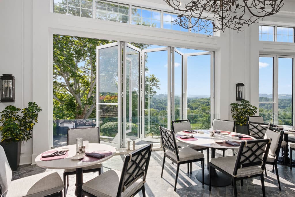 An elegant sunroom dining area featuring large, open white folding glass doors and high transom windows, showcasing a seamless Hurricane Impact Windows Installation overlooking a lush, scenic outdoor landscape.