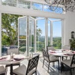 An elegant sunroom dining area featuring large, open white folding glass doors and high transom windows, showcasing a seamless Hurricane Impact Windows Installation overlooking a lush, scenic outdoor landscape.