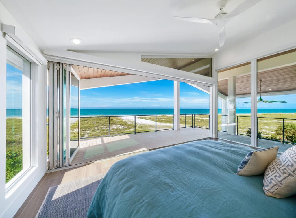 A bedroom view looking out past massive open folding glass doors onto an oceanfront balcony, featuring natural light from a complete Hurricane Impact Windows Installation.