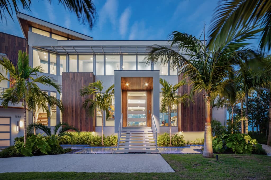 Exterior twilight view of a luxury modern home surrounded by palm trees, showcasing extensive floor-to-ceiling glass wall panels and an illuminated front entry from a custom Hurricane Impact Windows Installation.