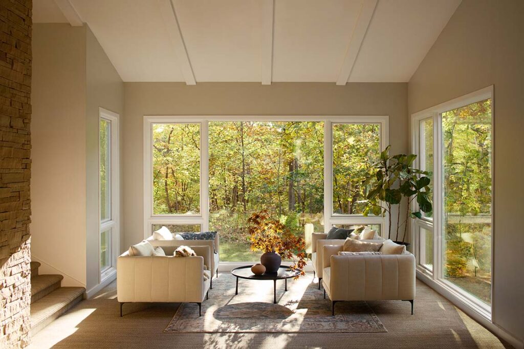 A sunlit, modern living room with plush armchairs and large picture replacement windows framing panoramic views of autumn foliage in a backyard setting.